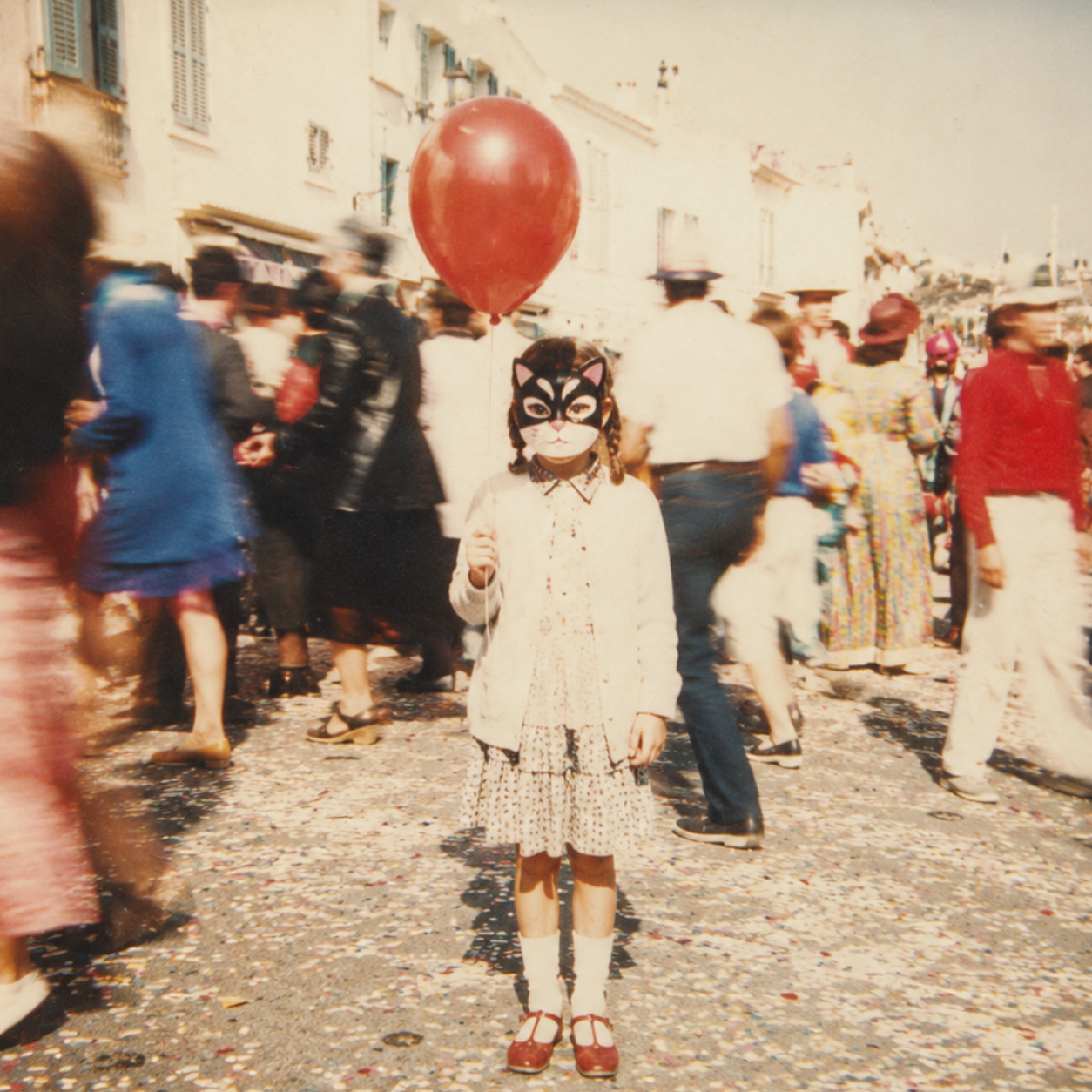 A child in a black cat mask holding a red balloon at a confetti-strewn street carnival